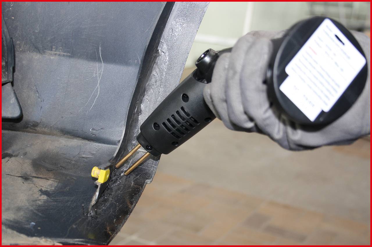 Eine Person trägt Handschuhe und hält ein Heizgerät, um Kunststoff an einem grauen Fahrzeugteil zu reparieren.