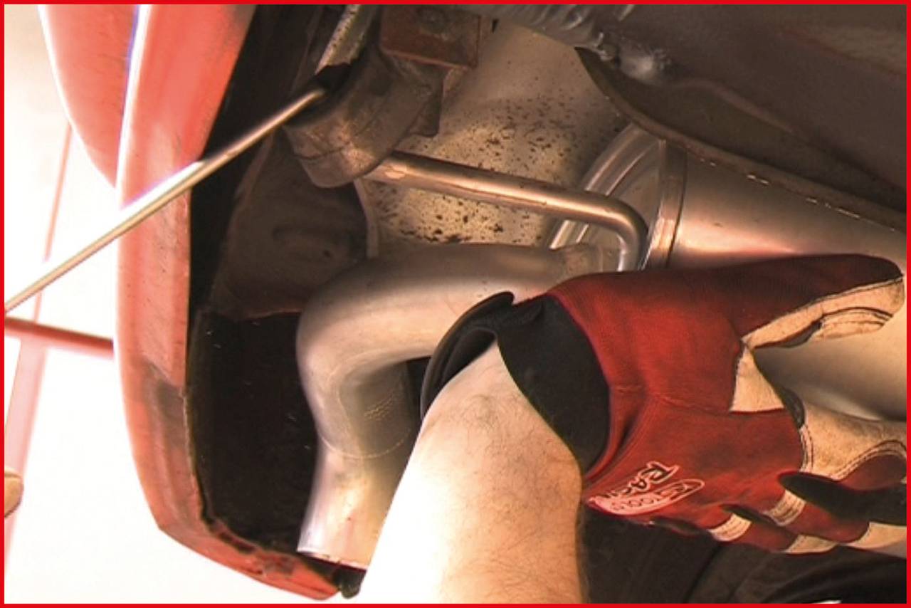 Ein Mechaniker mit roten Handschuhen arbeitet an einem Abgasrohr unter einem Auto. Im Hintergrund sind Teile des Auspuffs sichtbar.