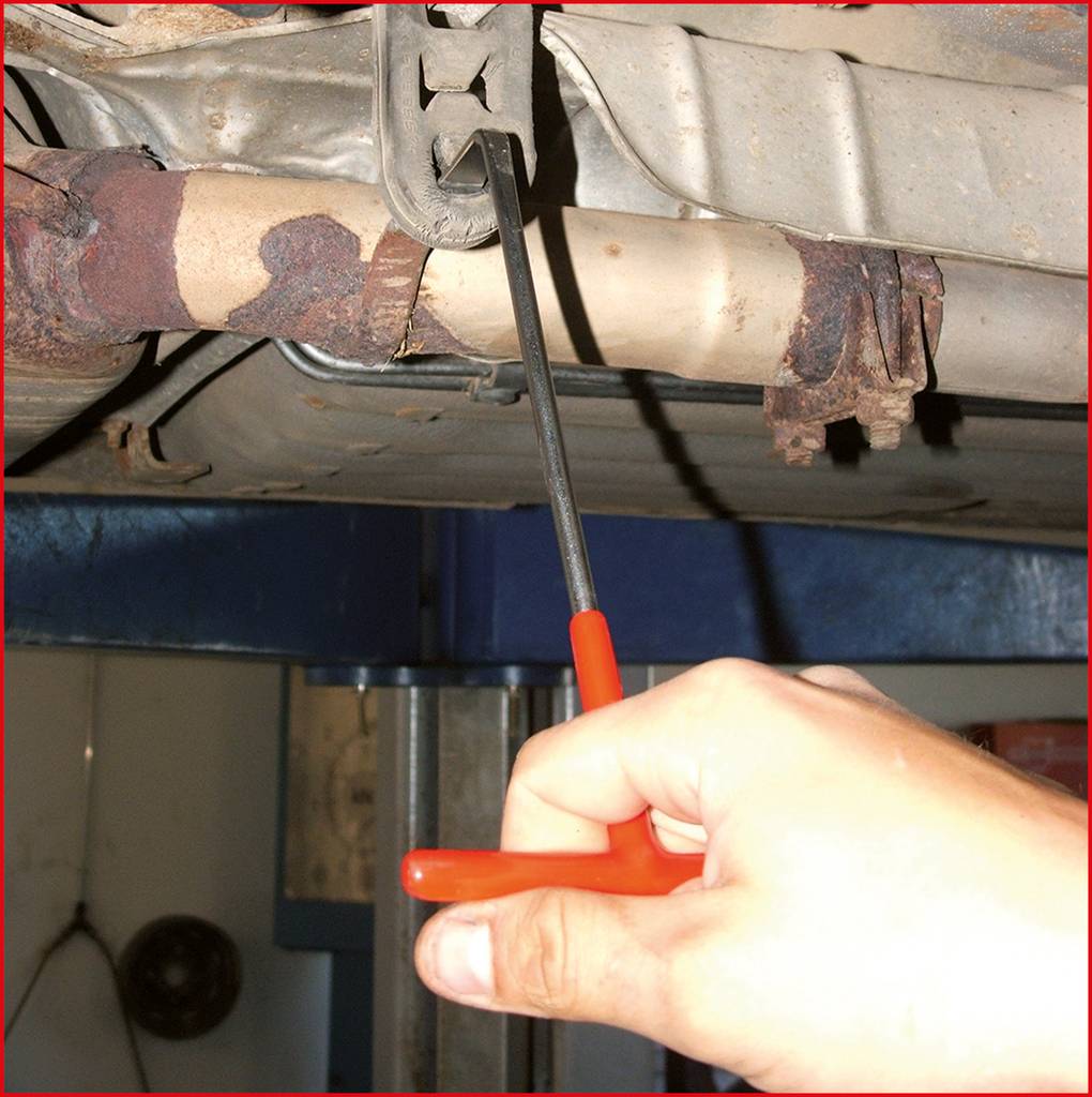 Eine Hand löst mit einem Schraubendreher eine Schraube an einem Autoauspuff, das auf einer Hebebühne in einer Werkstatt ist.