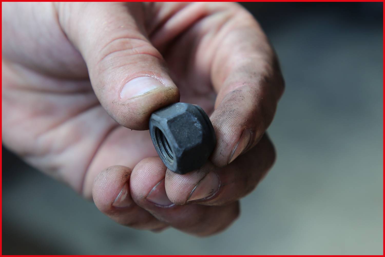 Eine Hand hält eine große, schwarze Sechskantmutter mit leichten Gebrauchsspuren. Die Finger sind leicht schmutzig.