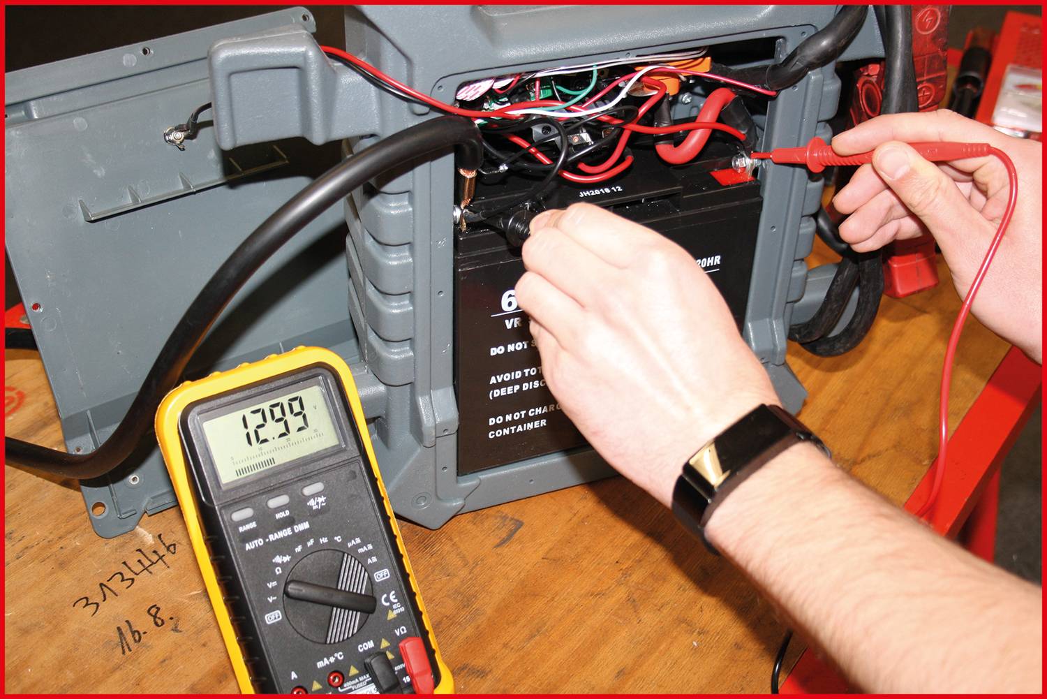 Eine Person misst die Spannung einer Batterie mit einem Multimeter. Das Display zeigt 12,99 Volt an. Kabel sind sichtbar verbunden.