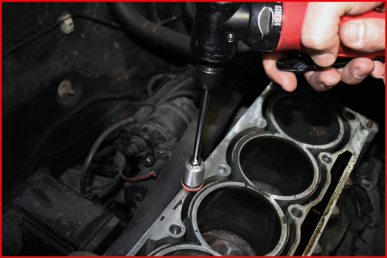 Eine Hand benutzt einen Elektrowerkzeug, um eine Schraube an einem Zylinderkopf eines Motors anzuziehen. Der Motorraum ist sichtbar.