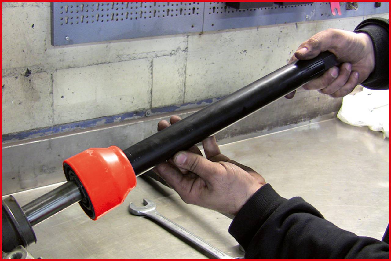 Eine Person hält ein schwarzes Metallrohr mit einer leuchtend orangefarbenen Schutzkappe in einer Werkstatt. Ein Schraubenschlüssel liegt daneben.