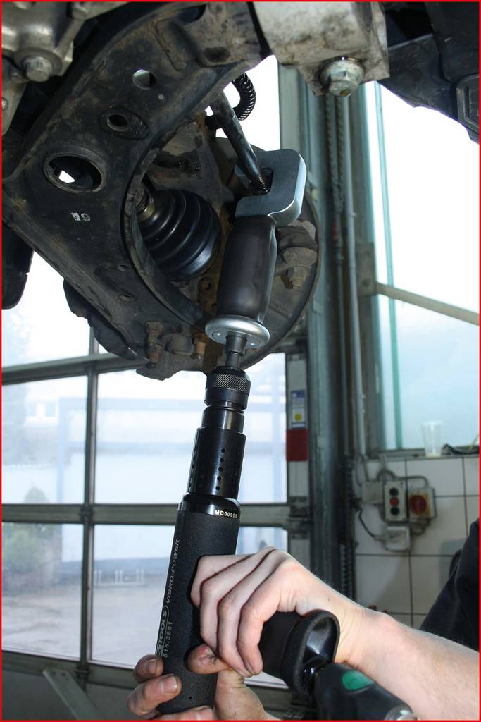 Ein Mechaniker verwendet ein Spezialwerkzeug, um eine Fahrzeugaufhängung zu reparieren. Die Werkstatt ist im Hintergrund sichtbar.