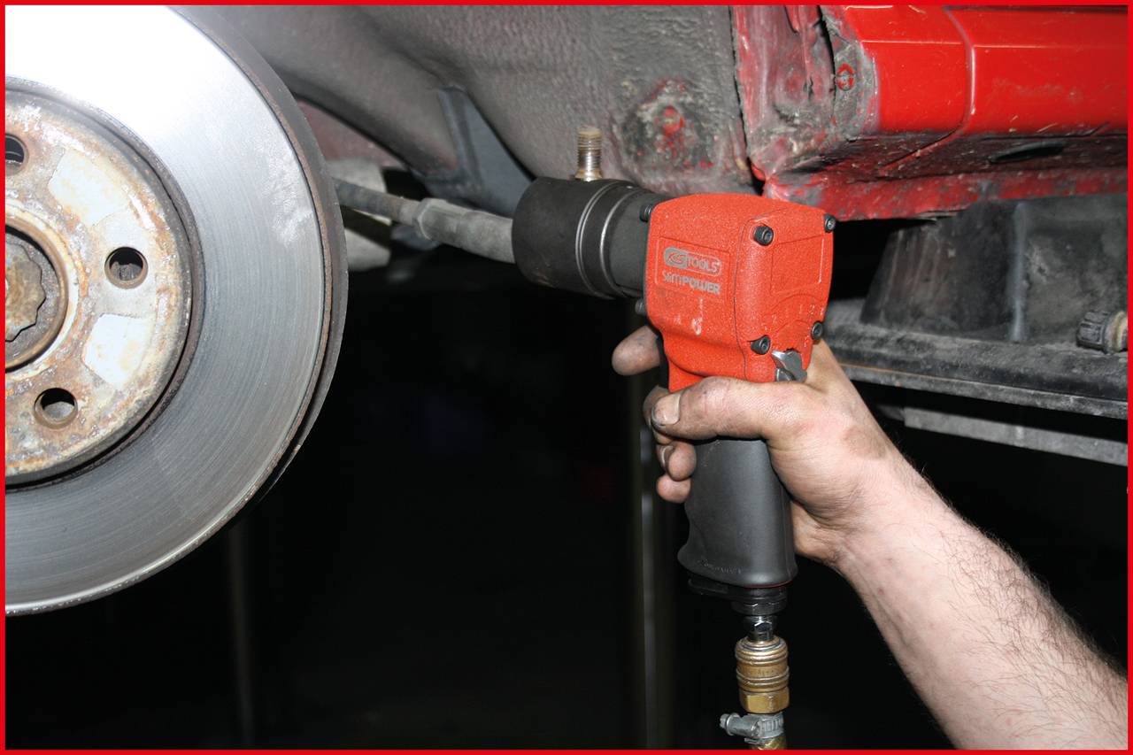 Ein Mechaniker benutzt einen Druckluftschrauber, um eine Radmutter an einem Auto zu lösen. Im Hintergrund ist die Bremsscheibe sichtbar.