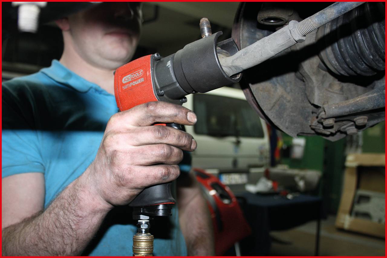 Ein Mechaniker verwendet ein Druckluftwerkzeug, um an einem Fahrzeugteil zu arbeiten. Im Hintergrund sind Werkzeuge und ein Auto zu sehen.