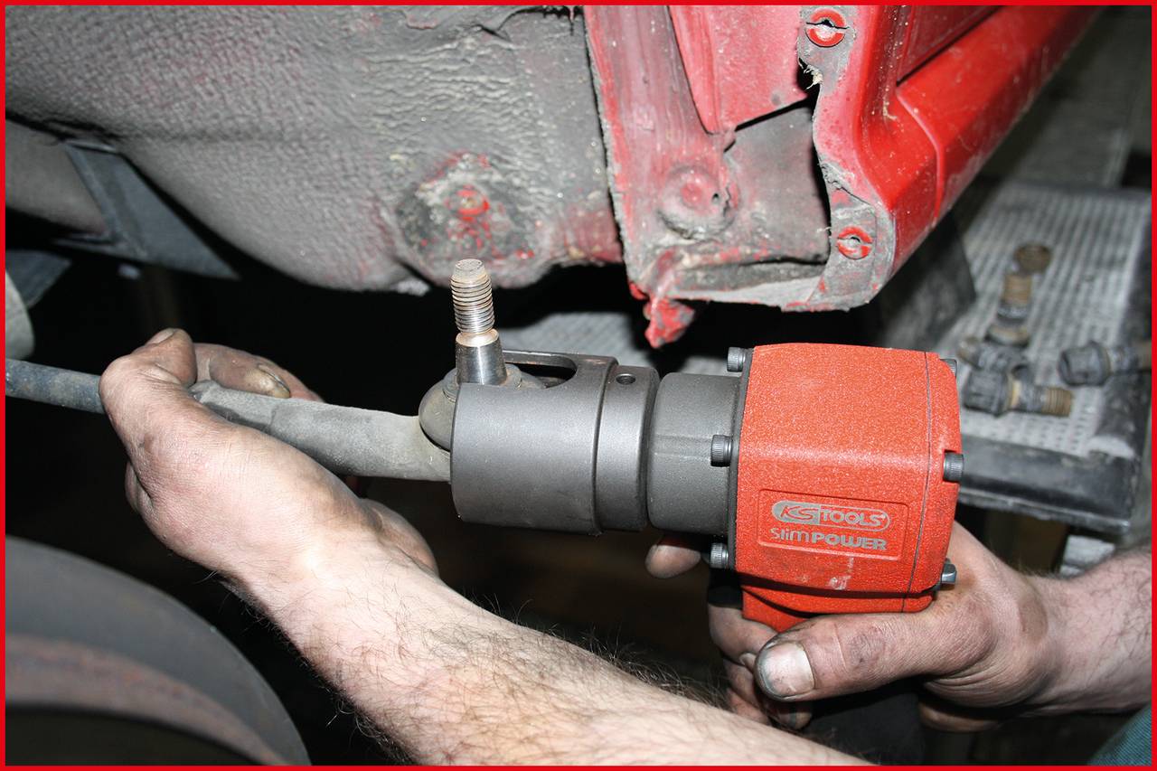 Nahaufnahme eines Mechanikers, der eine Achsmutter mit einem Druckluftwerkzeug löst. Staubige Fahrzeugunterseite im Hintergrund.