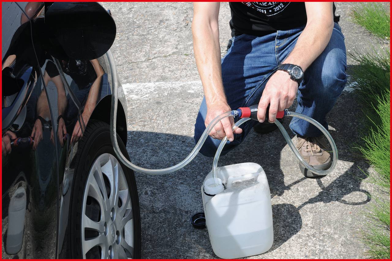 Person füllt mit einem Schlauch Benzin aus einem weißen Kanister in ein Auto. Outdoor-Szene, Straße und Gras im Hintergrund.