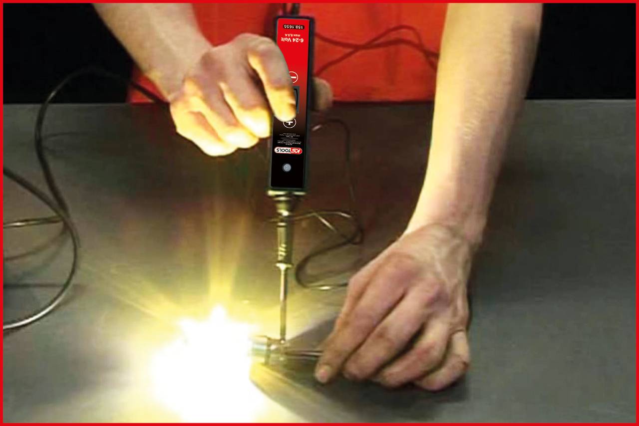 Person benutzt einen Lötkolben, um ein Metallteil mit einem leuchtenden Punkt zu löten. Die Umgebung ist dunkel, Fokus auf die Hände.