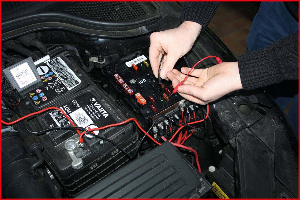 Zwei Hände arbeiten an der Autobatterie mit Kabeln und Sicherungen, umgeben von verschiedenen Autoteilen im Motorraum.