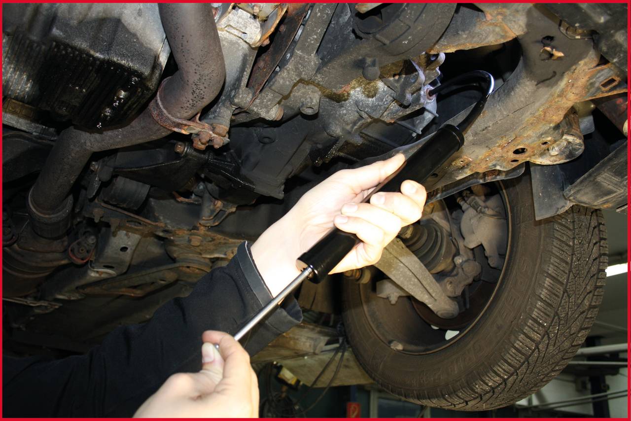 Mechaniker repariert den Unterboden eines Autos in einer Werkstatt. Die Person hält ein Werkzeug und arbeitet an der Fahrzeugaufhängung.