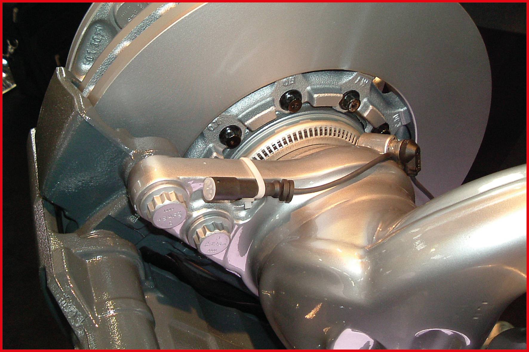 Nahaufnahme von einer silbernen Auto-Bremsscheibe mit Bremssattel, Kabeln und Befestigungsteilen in einer Werkstatt.