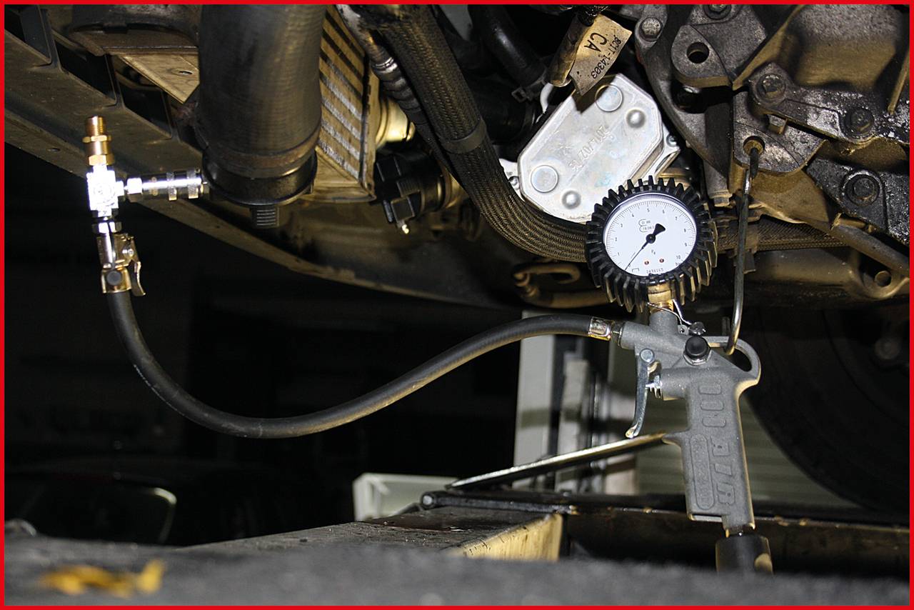 Nahaufnahme einer Fahrzeugunterseite während einer Druckprüfung. Ein Manometer ist an den Motor angeschlossen, um den Druck zu messen.