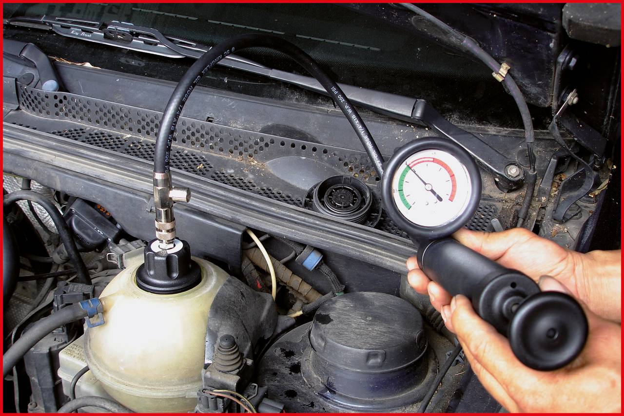 Messgerät zeigt Druck im Kühlkreislauf eines Autos an. Hände halten das Gerät, der Druck ist im roten Bereich. Motorraum im Hintergrund.
