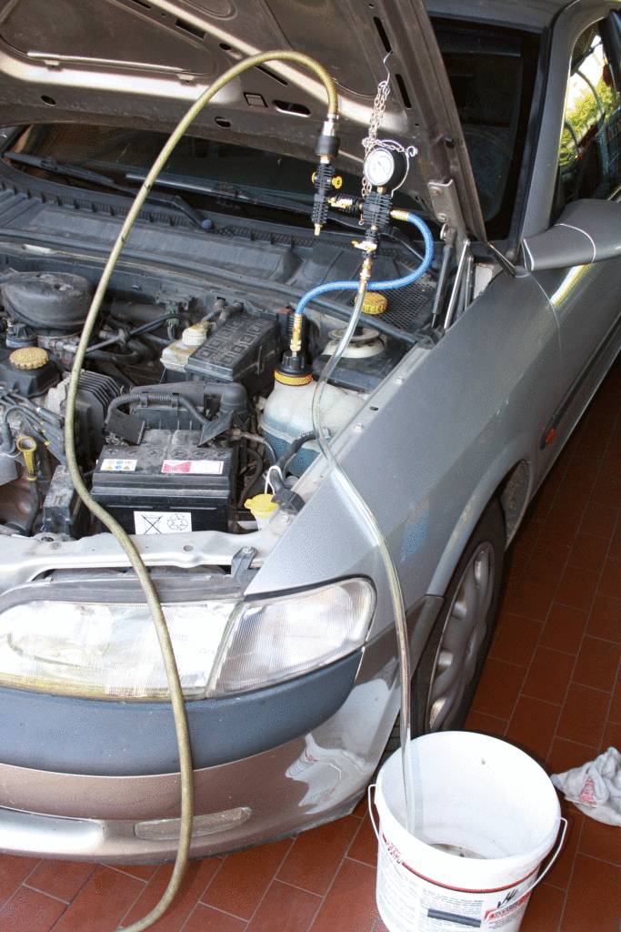 Ein Auto mit geöffneter Motorhaube wird entlüftet. Schläuche und ein Manometer sind am Motor angeschlossen, ein Eimer fängt Flüssigkeit auf.