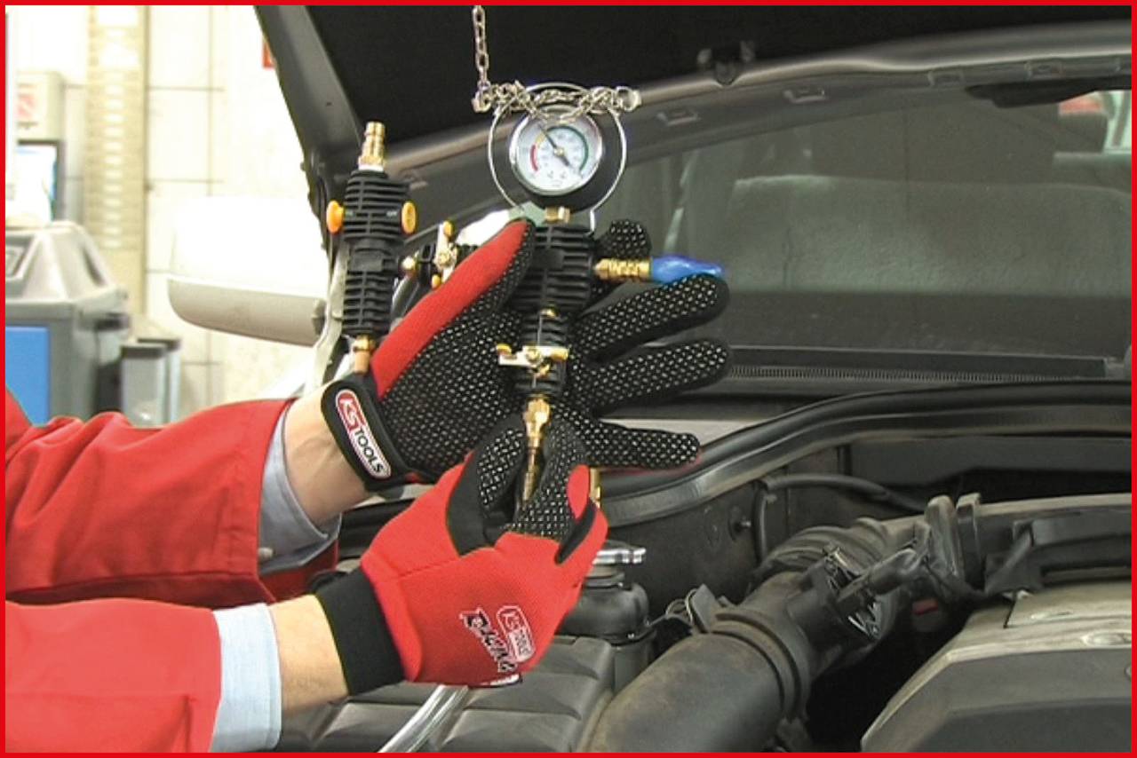 Eine Person in roten Handschuhen hält ein Manometer in der Nähe eines offenen Motorraums, möglicherweise zur Druckmessung.