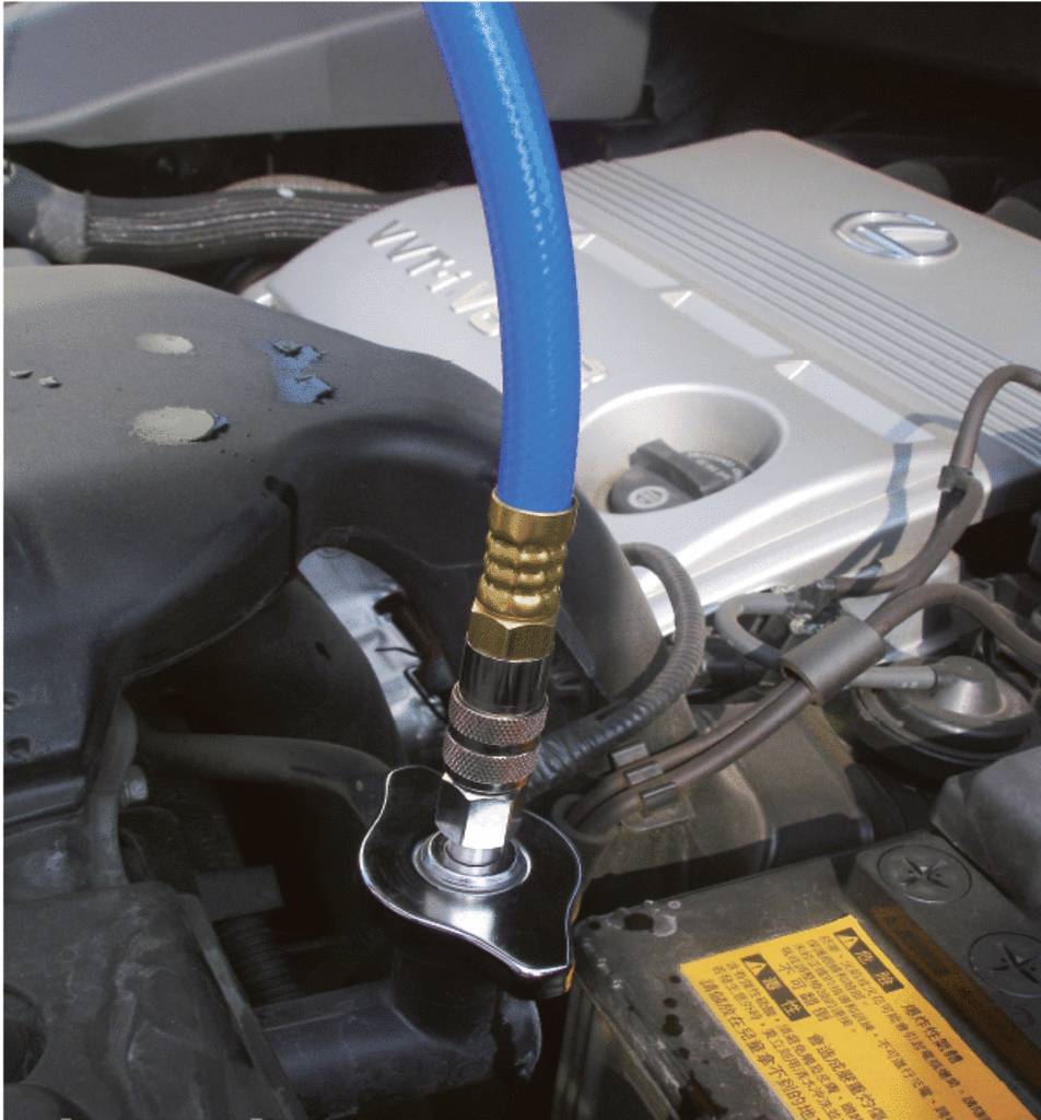 Ein blauer Schlauch ist an den Kühlmittelbehälter eines Autokompressors angeschlossen. Hintergrund zeigt Motorteile.