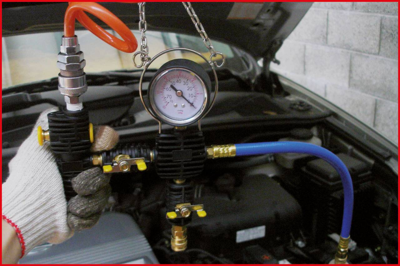 Ein Mechaniker hält ein Druckmessgerät mit Schläuchen in der Hand vor einem geöffneten Auto. Das Auto befindet sich in einer Werkstatt.