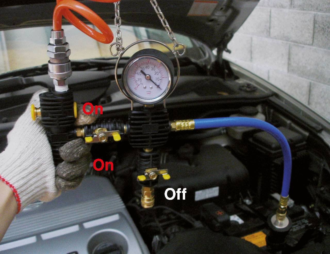 Ein Manometer zeigt den Druck in einer Autowerkstatt. Zwei Ventile sind mit 'On', ein Ventil mit 'Off' markiert. Motorhaube offen.