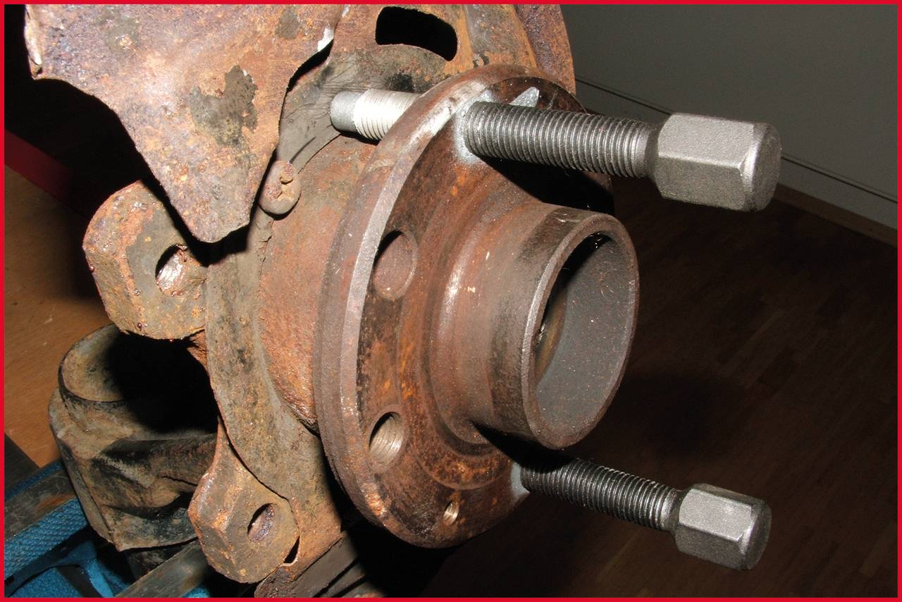 Ein rostiges Autoteil mit zwei langen Schrauben in einem Metallradlager. Das Teil liegt auf einer Holzoberfläche.