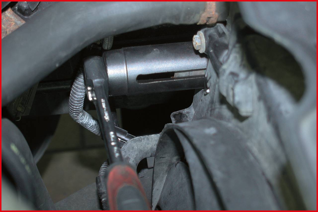 Ein Mechanikerwerkzeug greift einen runden, metallischen Teil in einem Motorraum. Es werden Reparaturen oder Anpassungen durchgeführt.