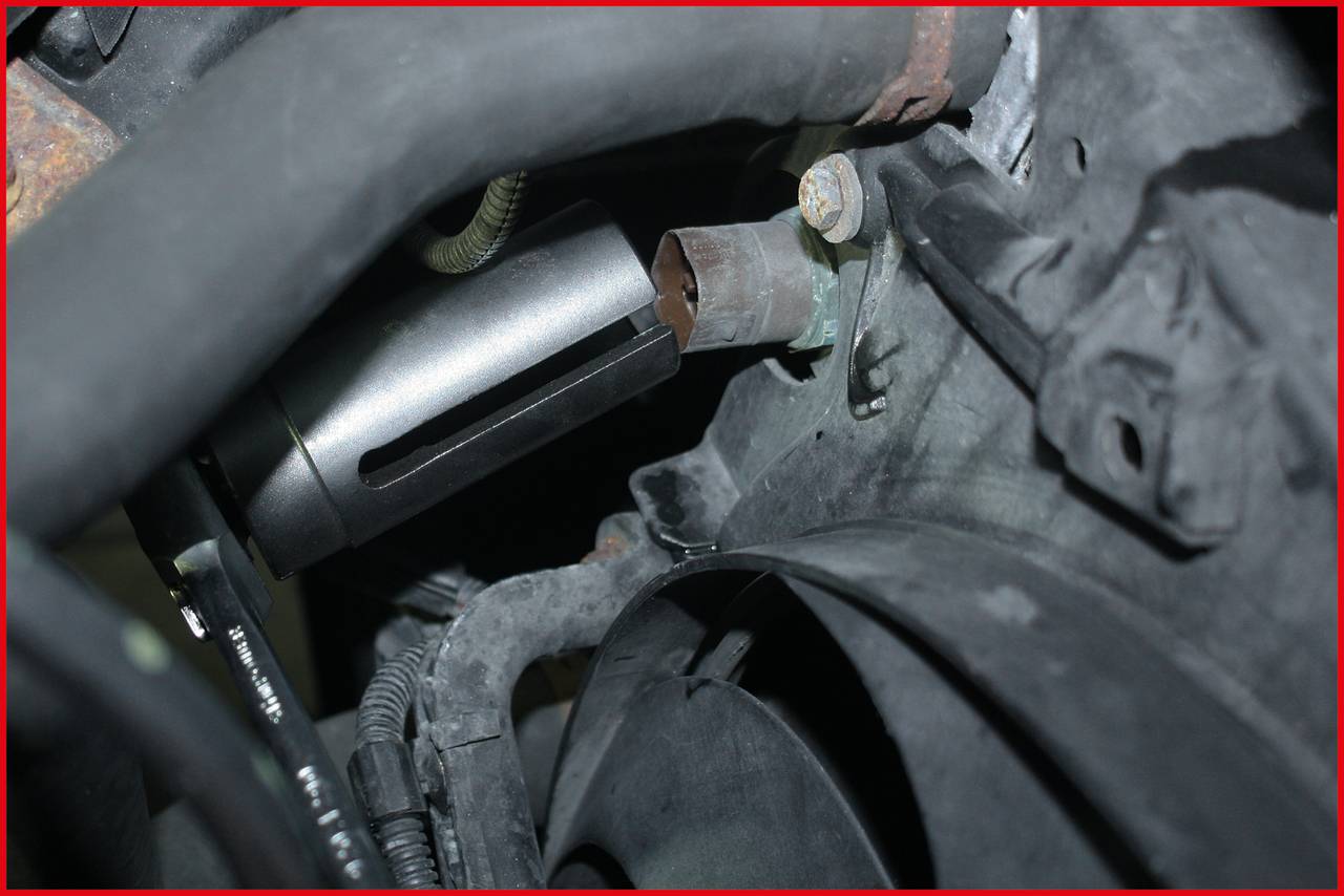 Ein Bauteil in einem Motorraum zeigt ein Metallzylinder an einer Halterung, umgeben von Schläuchen und Kabeln.