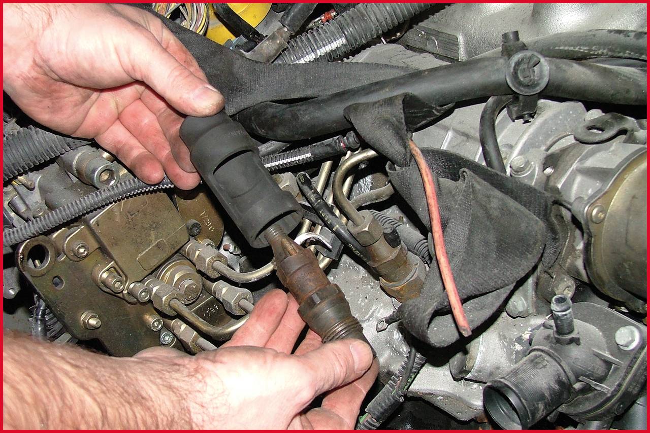 Eine Person hält ein Autoteil, möglicherweise eine Zündkerze, nahe dem Motor eines Fahrzeugs. Kabel und Schläuche sind sichtbar.