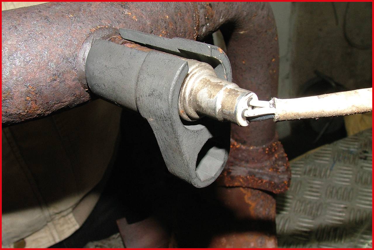 Eine Zündkerze ist in einen Auspuff eingebaut. Sie ist durch Rost und Verschleiß gekennzeichnet und dient vermutlich zur Funktionsprüfung.