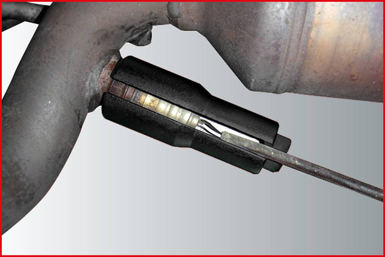 Ein schwarzes Bauteil ist an einem Rohr befestigt, vermutlich Teil eines Automotor- oder Abgassystems. Ein Kabel ist angeschlossen.