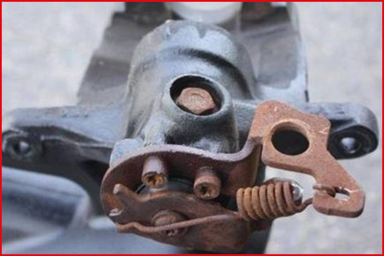 Ein rostiges mechanisches Teil mit Schrauben und Feder auf grauem Hintergrund, möglicherweise Teil eines Maschinenmotors oder Fahrzeugs.