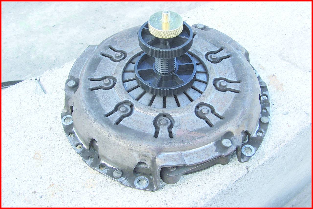 Eine Kupplungsscheibe mit Druckplatte auf einem Betonuntergrund, zentraler Gewindebolzen und schwarze, radförmige Struktur sichtbar.