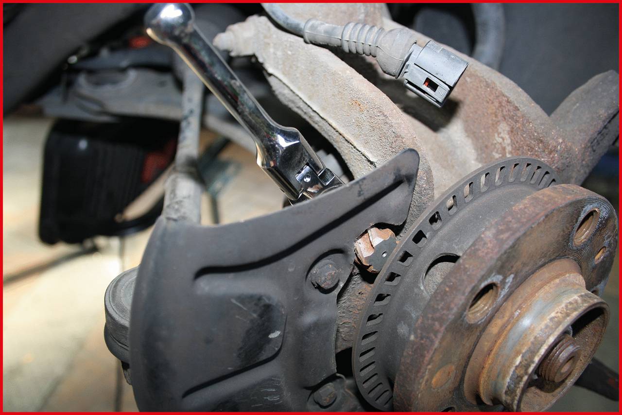 Nahaufnahme einer Autoreparatur: Eine Ratsche zieht eine Schraube an einer Fahrzeugaufhängung fest. Rostige Bremskomponente im Hintergrund.