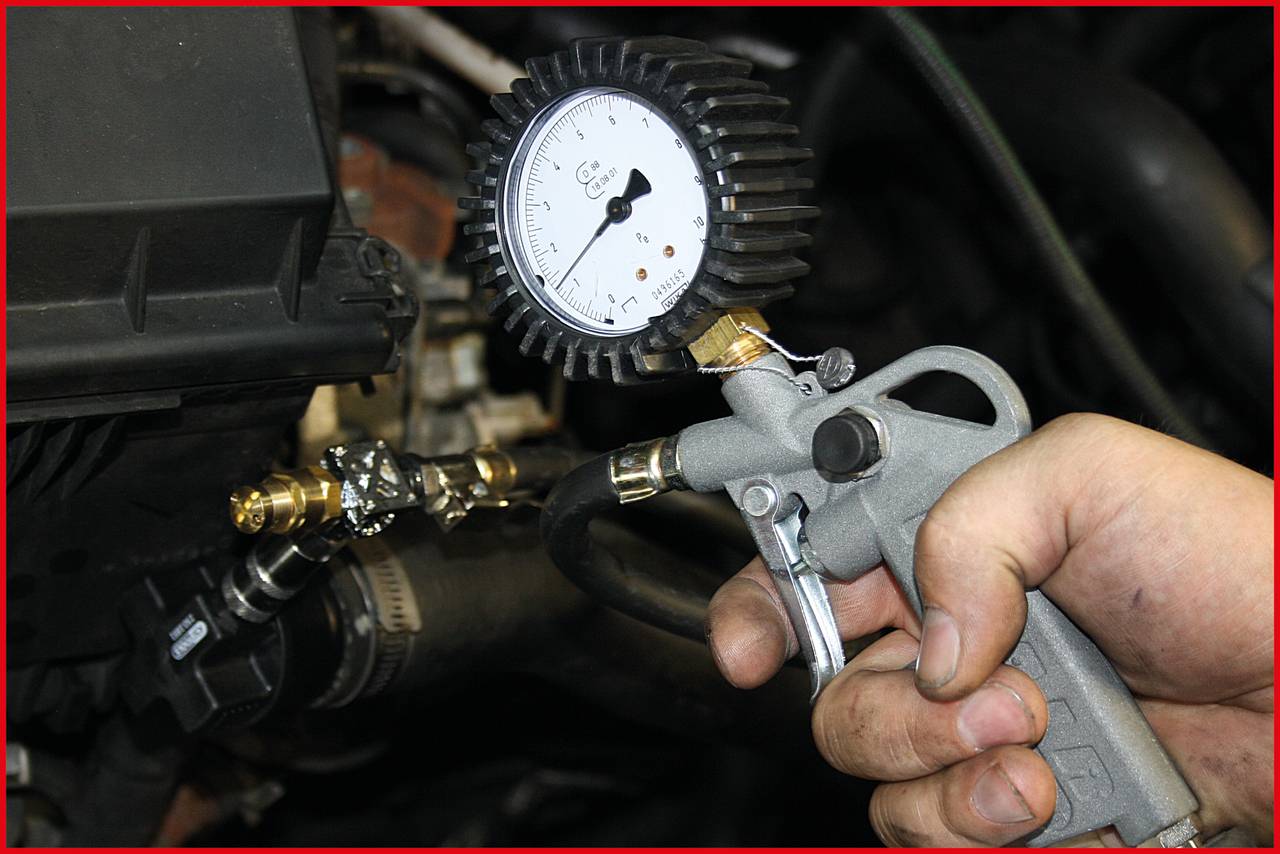 Eine Hand hält einen Reifendruckmesser mit einem Manometer, das mit einem Schlauch verbunden ist. Hintergrund zeigt Motorteile.