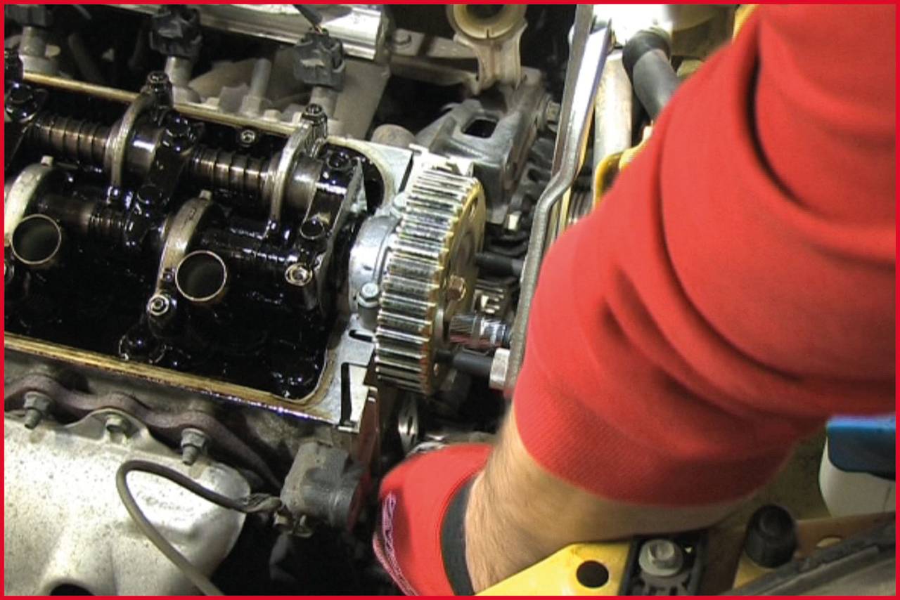 Ein Mechaniker in roter Kleidung repariert einen offenen Automotor. Zu sehen sind Zahnräder und Motorteile in Nahaufnahme.