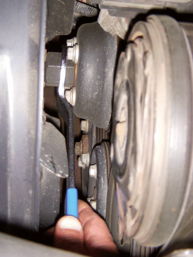 Eine Hand hält einen blauen Schraubenschlüssel, der in der Nähe eines Autoteils arbeitet, möglicherweise an einem Keilriemen oder einer Riemenscheibe.