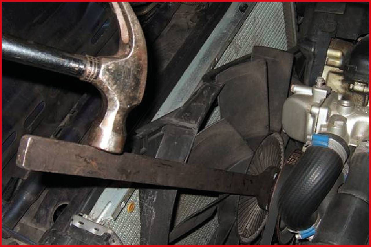 Ein Hammer schlägt auf einen Metallstab, der an einem Autokühler fixiert zu sein scheint. Ein Lüfter und Kühlerschläuche sind sichtbar.