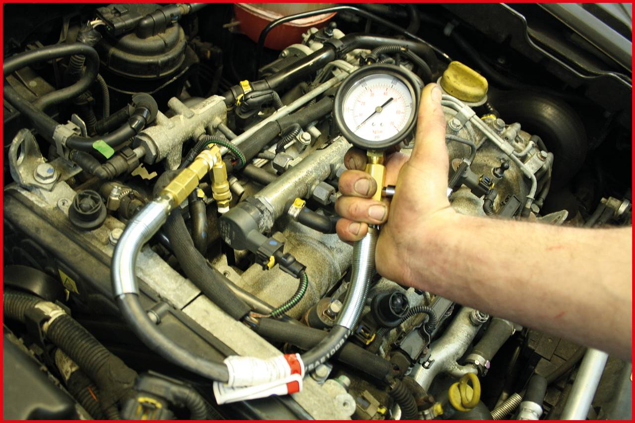 Eine Hand hält einen Druckmesser über einen Motor, um den Kompressionsdruck zu überprüfen. Es werden Schläuche und Motorteile sichtbar.
