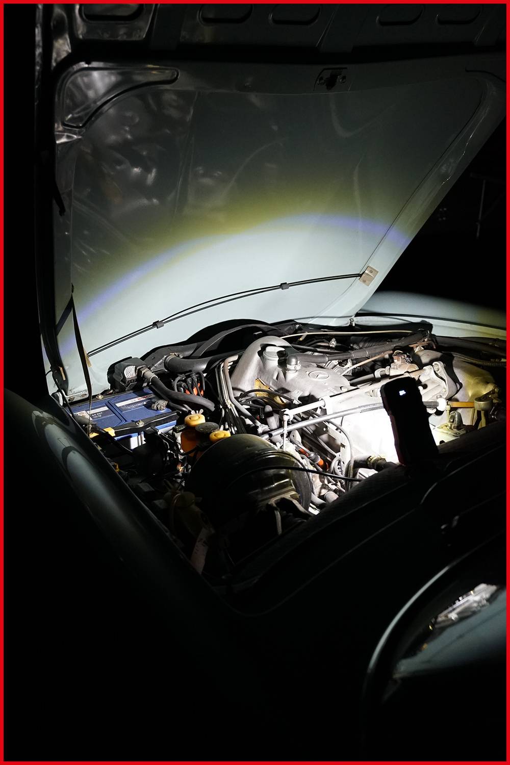 Motorraum eines Autos bei geöffneter Motorhaube, beleuchtet von Taschenlampe, zeigt Batterie und Motorteile im Detail.
