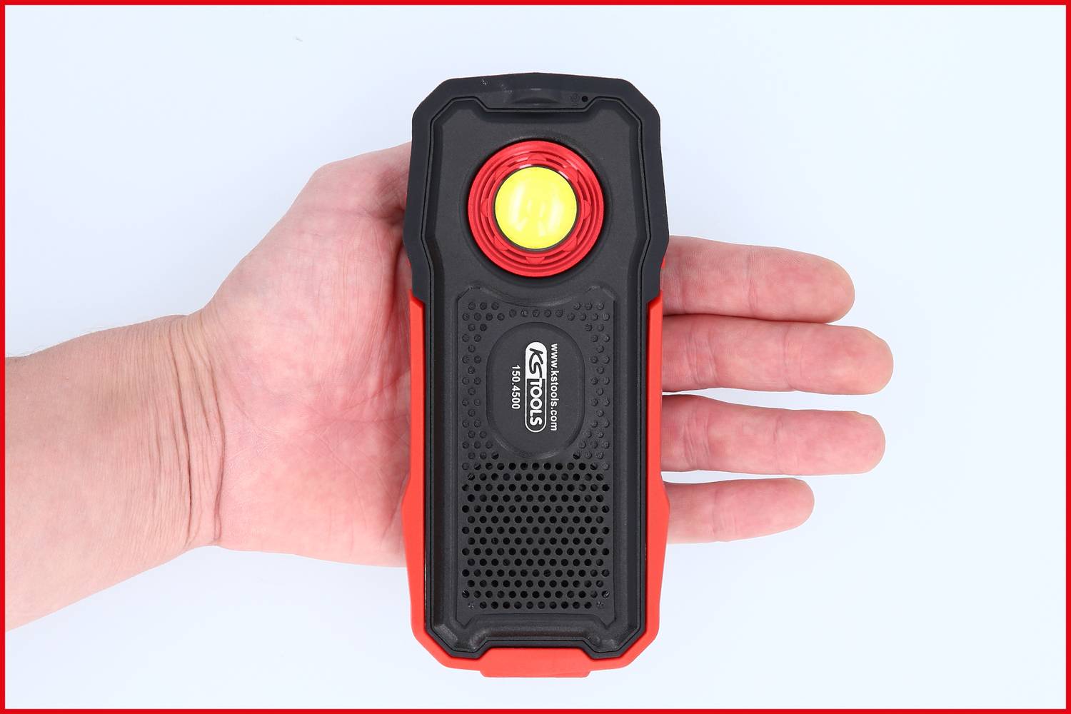 Eine Hand hält eine aufgeladene, rechteckige LED-Arbeitsleuchte mit markantem rotem Rand und schwarzer Vorderseite mit gelbem Licht.