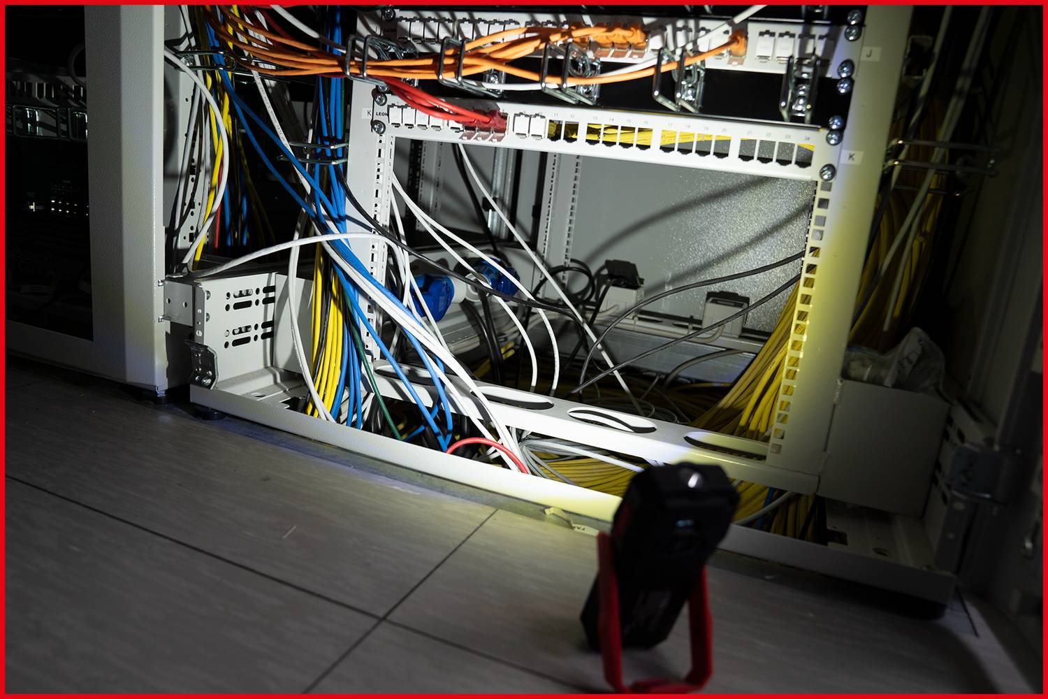 Ein offener Netzwerkschrank mit vielen Kabeln in verschiedenen Farben. Im Vordergrund leuchtet eine Lampe den Schrank aus.