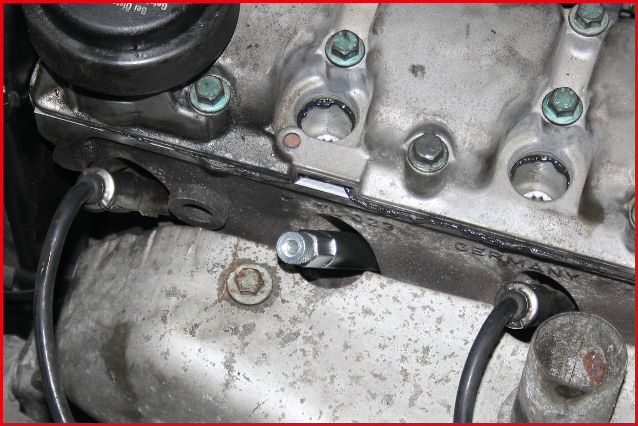 Motorteil mit Zündkerzen und Kabeln, verölte Oberfläche. Details von Schrauben und metallischer Struktur sind sichtbar.