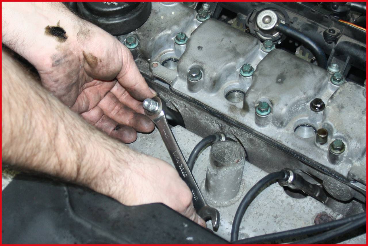 Eine Person repariert einen Automotor und hält einen Schraubenschlüssel an einer Schraube am Motorblock. Ölflecken sichtbar.