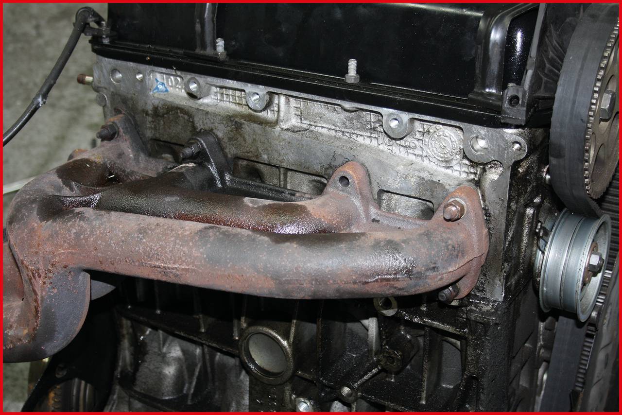 Ein verrosteter Auspuffkrümmer an einem Motorblock in einer Werkstattumgebung.