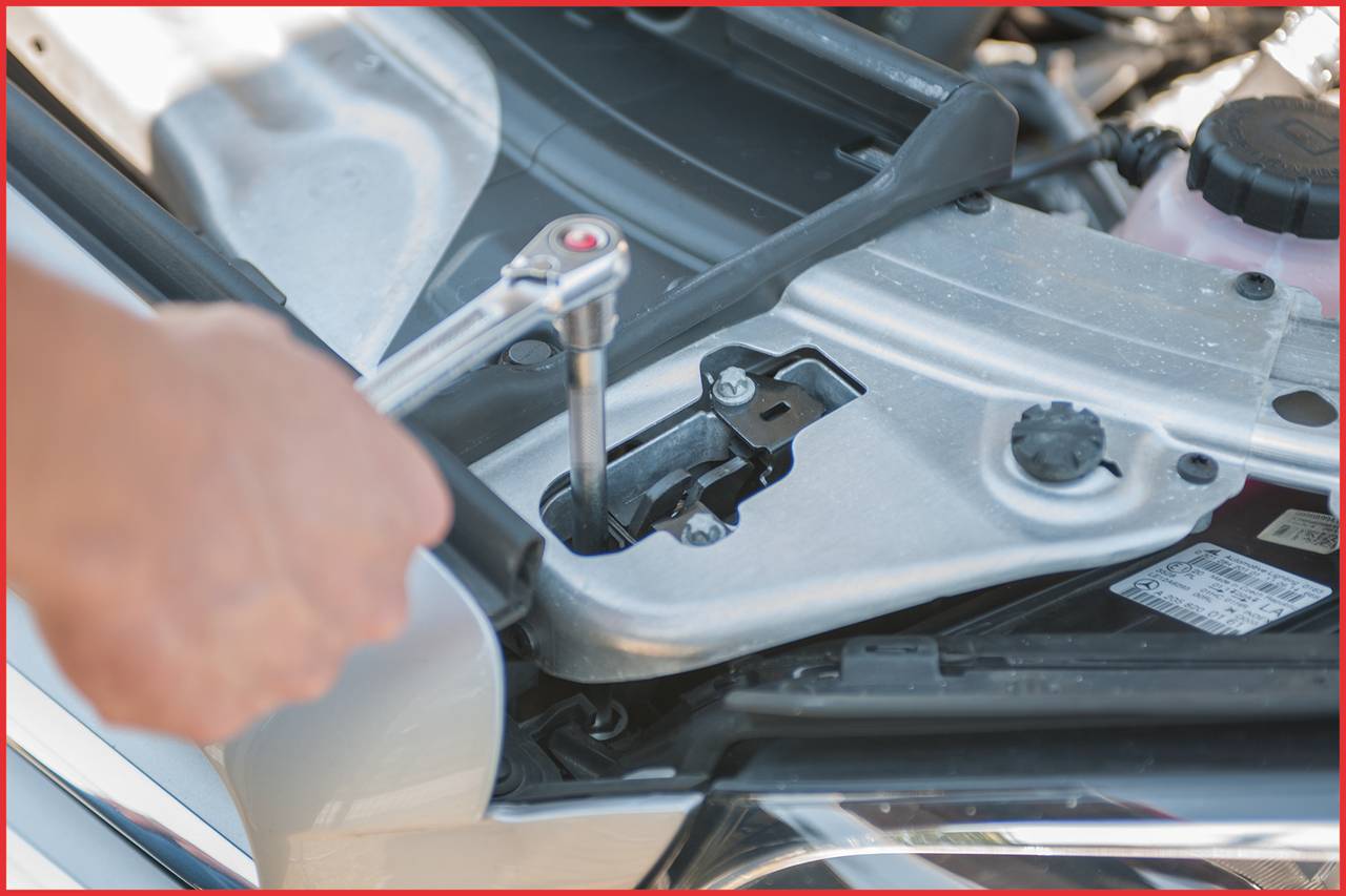 Eine Person zieht mit einem Schraubenschlüssel eine Schraube im Motorraum eines Autos an. Werkzeug und Autoteile sichtbar.
