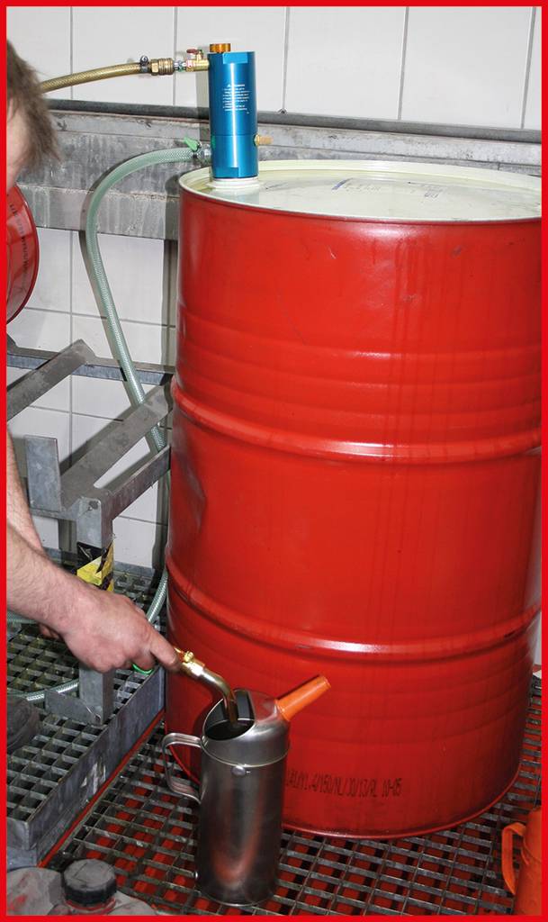 Ein rotes Metallfass mit Flüssigkeit. Eine Person füllt mit einem Zapfhahn einen silbernen Behälter vor einem industriellen Hintergrund.