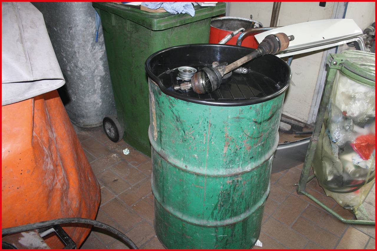 Ein grünes Metallfass steht in einer Werkstatt. Darauf liegt eine Autoantriebswelle. Im Hintergrund sind Mülltonnen und diverse Werkstattgegenstände zu sehen.