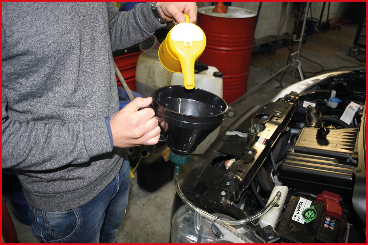 Eine Person schüttet Motoröl mit einem gelben Kanister über einen Trichter in ein Auto. Im Hintergrund sind Ölfässer zu sehen.