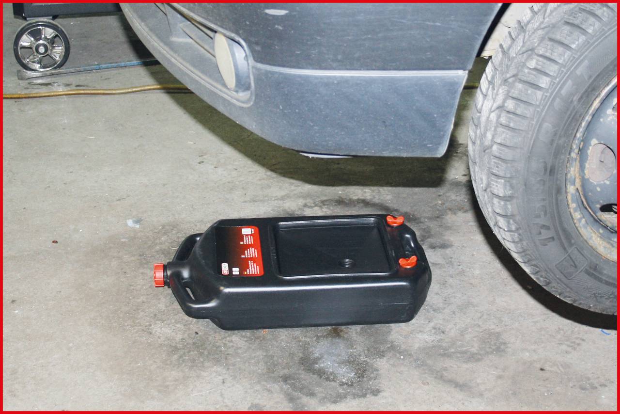 Ein schwarzer Ölauffangbehälter mit rotem Deckel steht unter dem Motorraum eines Autos in einer Garage. Links ist ein Wagenheber erkennbar.