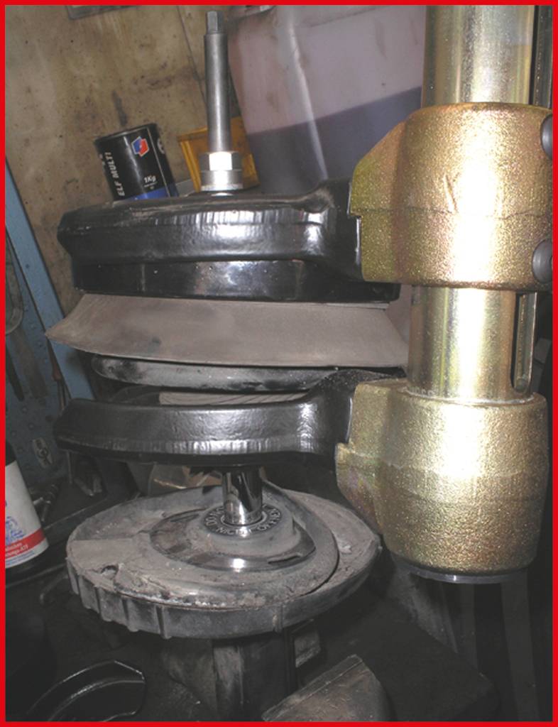 Fahrzeugfeder wird mit hydraulischer Maschine zusammengedrückt. Werkstattumgebung mit Werkzeugen im Hintergrund sichtbar.