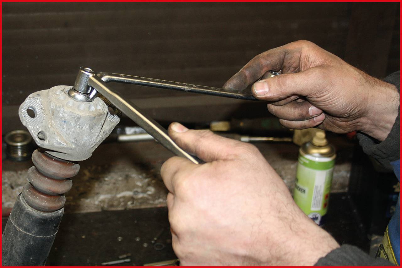 Hände einer Person reparieren eine Autoteil, Verwendung von zwei Schraubenschlüsseln, Arbeitstisch mit Werkzeugen im Hintergrund.